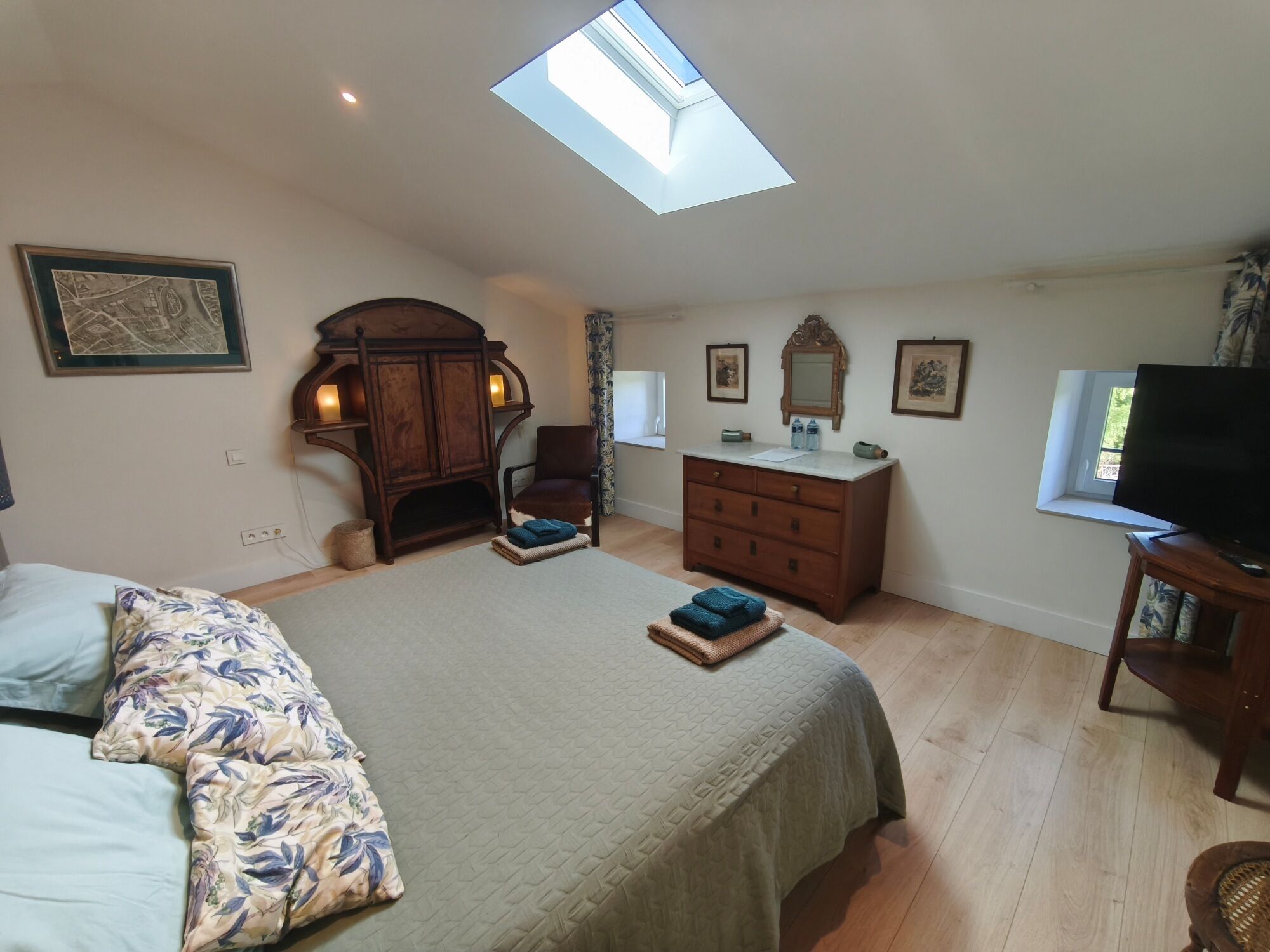 A bedroom with a skylight, a bed with floral bedding, a dresser, a chair, a television, and framed artwork on the walls.