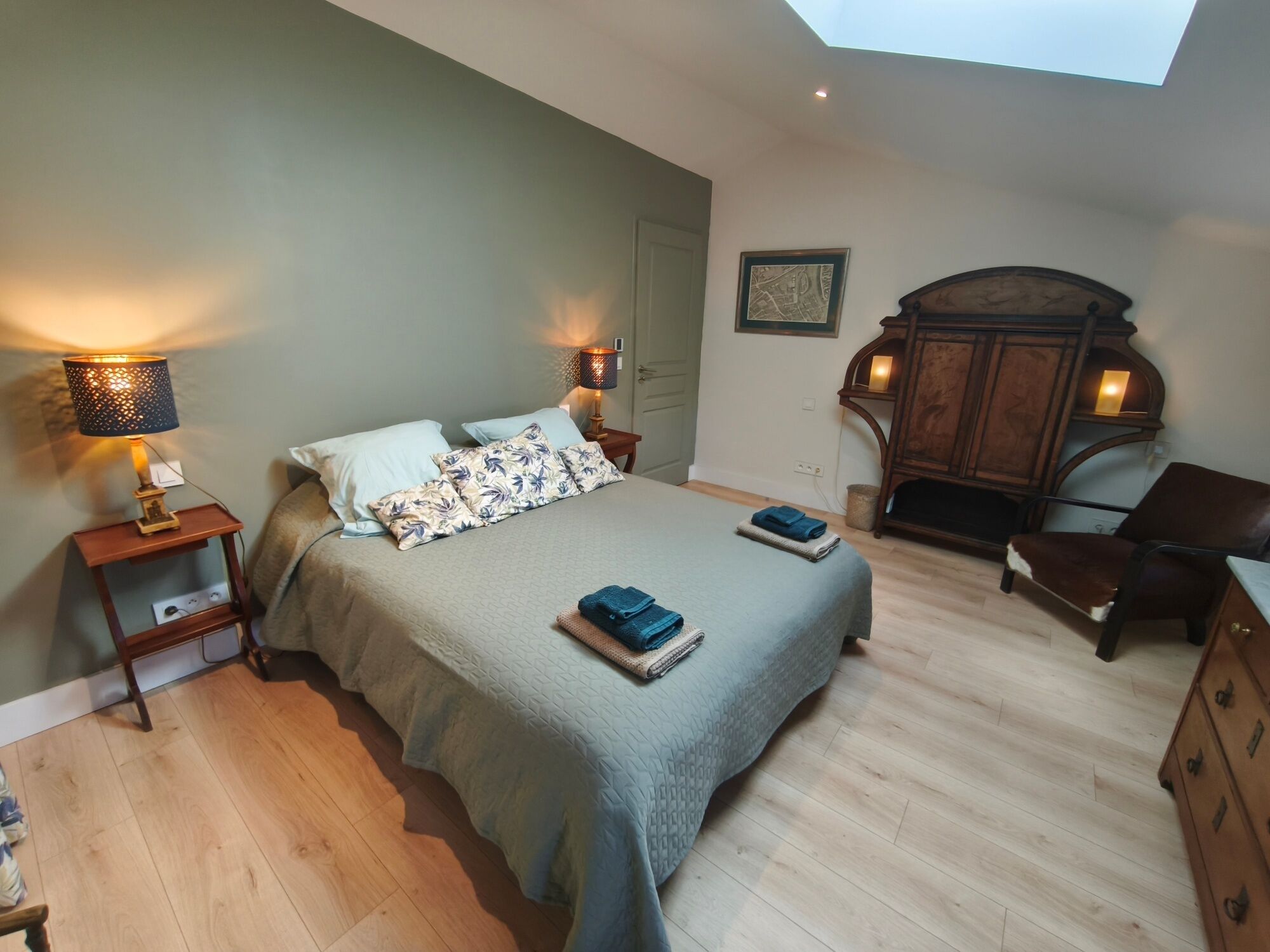 A bedroom with a bed, two bedside lamps, a wooden dresser, a chair, and a skylight.