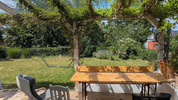An outdoor patio with a wooden table and chairs, surrounded by lush greenery and a view of a building in the background. - Vacation apartment Ludwigshöhe (Weißenburg)