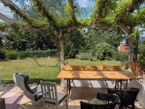 An outdoor patio with a wooden table and chairs, surrounded by lush greenery and a view of a building in the background. - Vacation apartment Ludwigshöhe (Weißenburg)