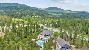 A residential area with a large house featuring a swimming pool, surrounded by dense forest and hills in the background. - Cabin · Hot Tub · Arcade · Trails · Sleeps 16 (Lead)