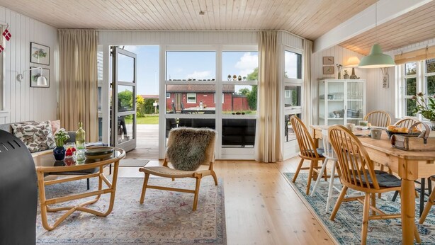 A spacious living room with a wooden ceiling, a dining area with a wooden table and chairs, and a large window offering a view of the outside.