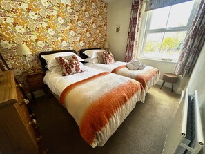 Habitación de hotel con dos camas, papel tapiz floral, una ventana con cortinas, un taburete pequeño y un radiador.
