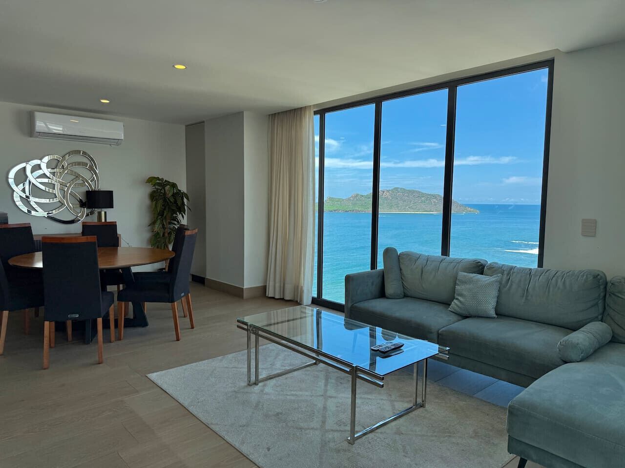 A modern living room with a large window, a sofa, a glass coffee table, and a dining area with chairs and a table.