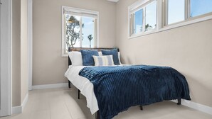 A bedroom with a bed, two windows, and a view of trees and a palm tree.