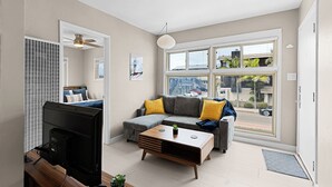 A modern living room with a sofa, a coffee table, and a TV. There are windows with a view of the outside.