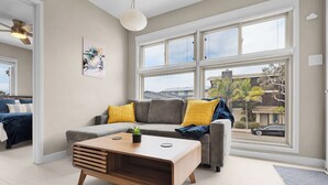 A modern living room with a grey sofa, yellow cushions, a wooden coffee table, and large windows offering a view of the street and palm trees.