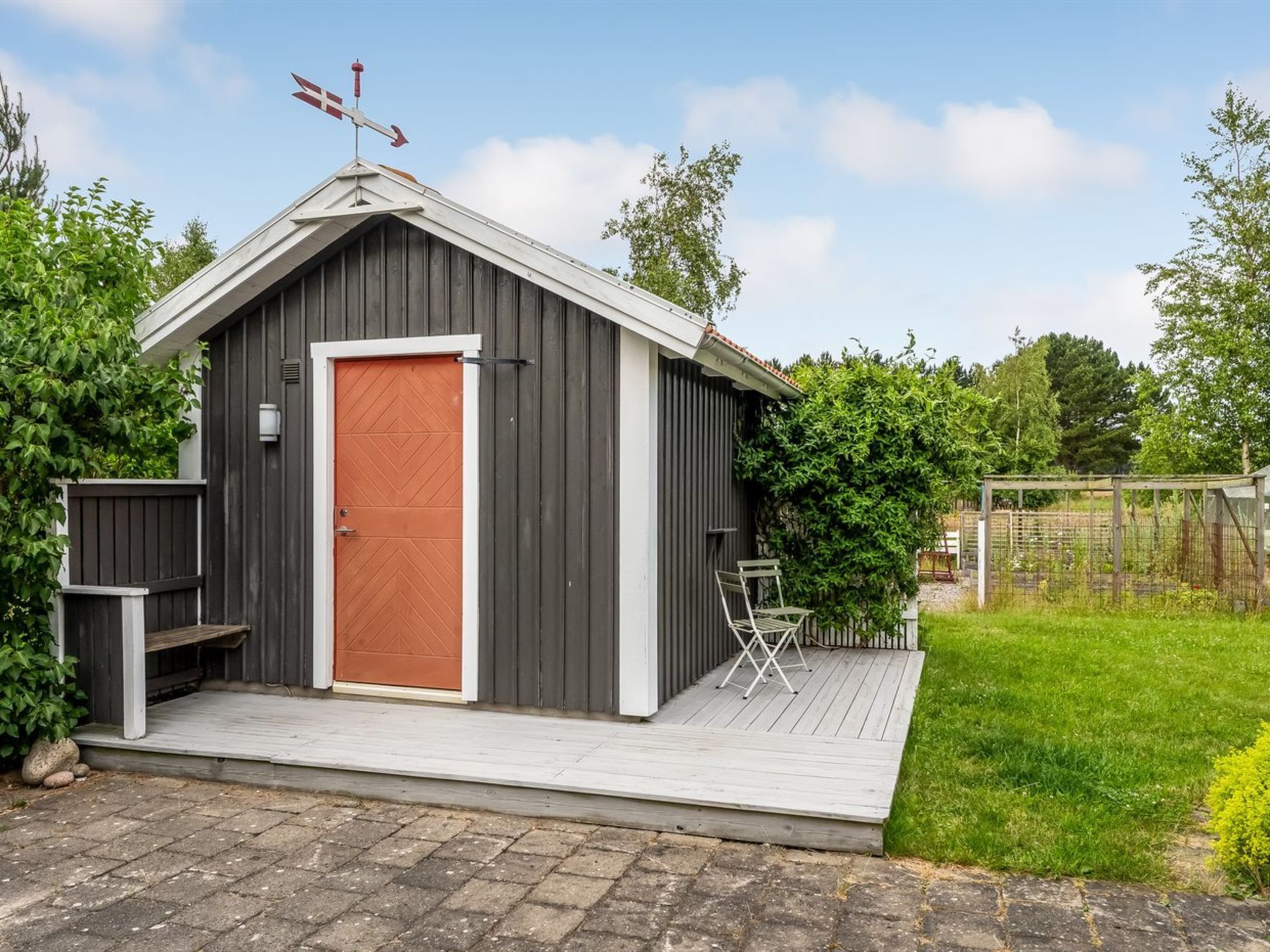 A small, dark wooden shed with a red door, a small porch, and a metal fence in the background.