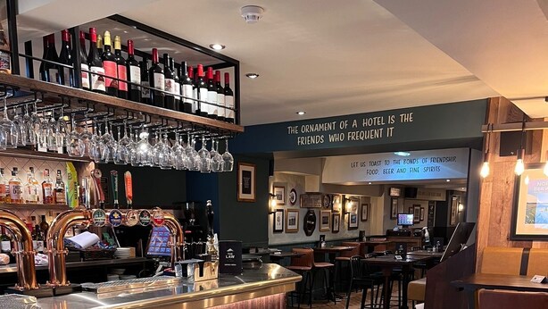 A bar area with a wooden counter, wine bottles, and glasses on shelves. There are tables and chairs in the background. - The Law (North Berwick)