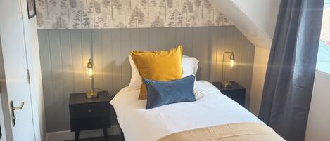 A bedroom with a sloped ceiling, a bed with a white duvet and two pillows, a bedside table with a lamp, and a patterned wallpaper.