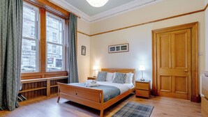 A neatly arranged bedroom with a wooden bed, a bedside table with a lamp, a window with curtains, and a door. - beautiful- spacious ground floor newington flat (edinburgh)