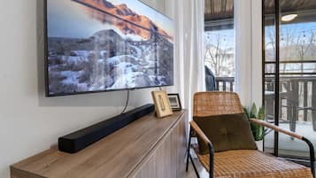 A room with a large flat-screen TV mounted on the wall, a wooden console table, a wicker chair, and a view through a large window.