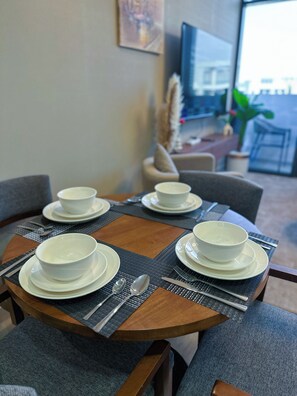 Una mesa de comedor con cuencos y platos blancos, un televisor al fondo y vista de la ciudad a través de una ventana.