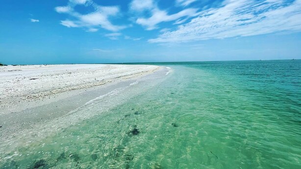 Una spiaggia con acque cristalline turchesi e una riva sabbiosa sotto un cielo azzurro intenso con nuvole sparse.