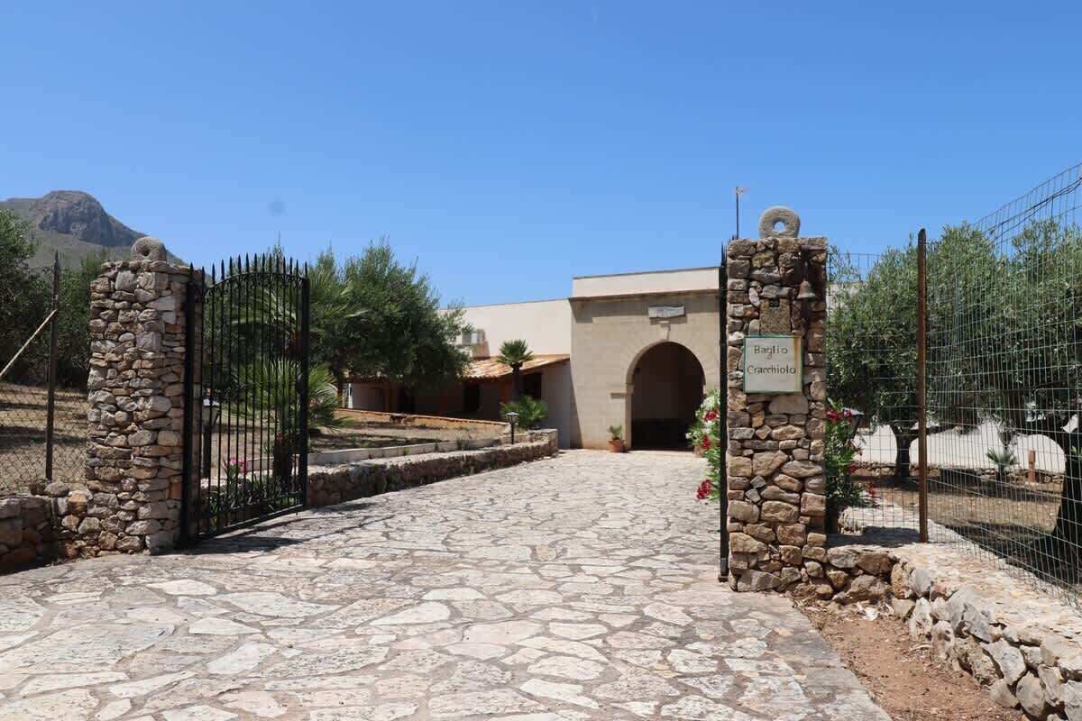 A stone-paved entrance with a gate, a building with an arched doorway, and a sign indicating 'Baglio Cacciolo.'