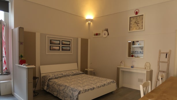 Een slaapkamer met een bed, nachtkastje, bureau, stoel en een klein plankje met een klok en foto’s.