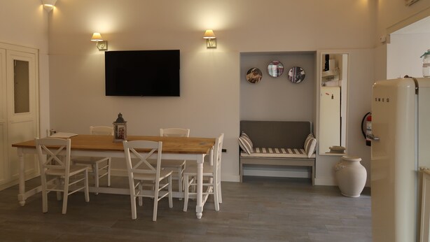 Ein Essbereich mit Holztisch und weißen Stühlen, einem an der Wand befestigten Flachbildfernseher und zwei Wandleuchten.