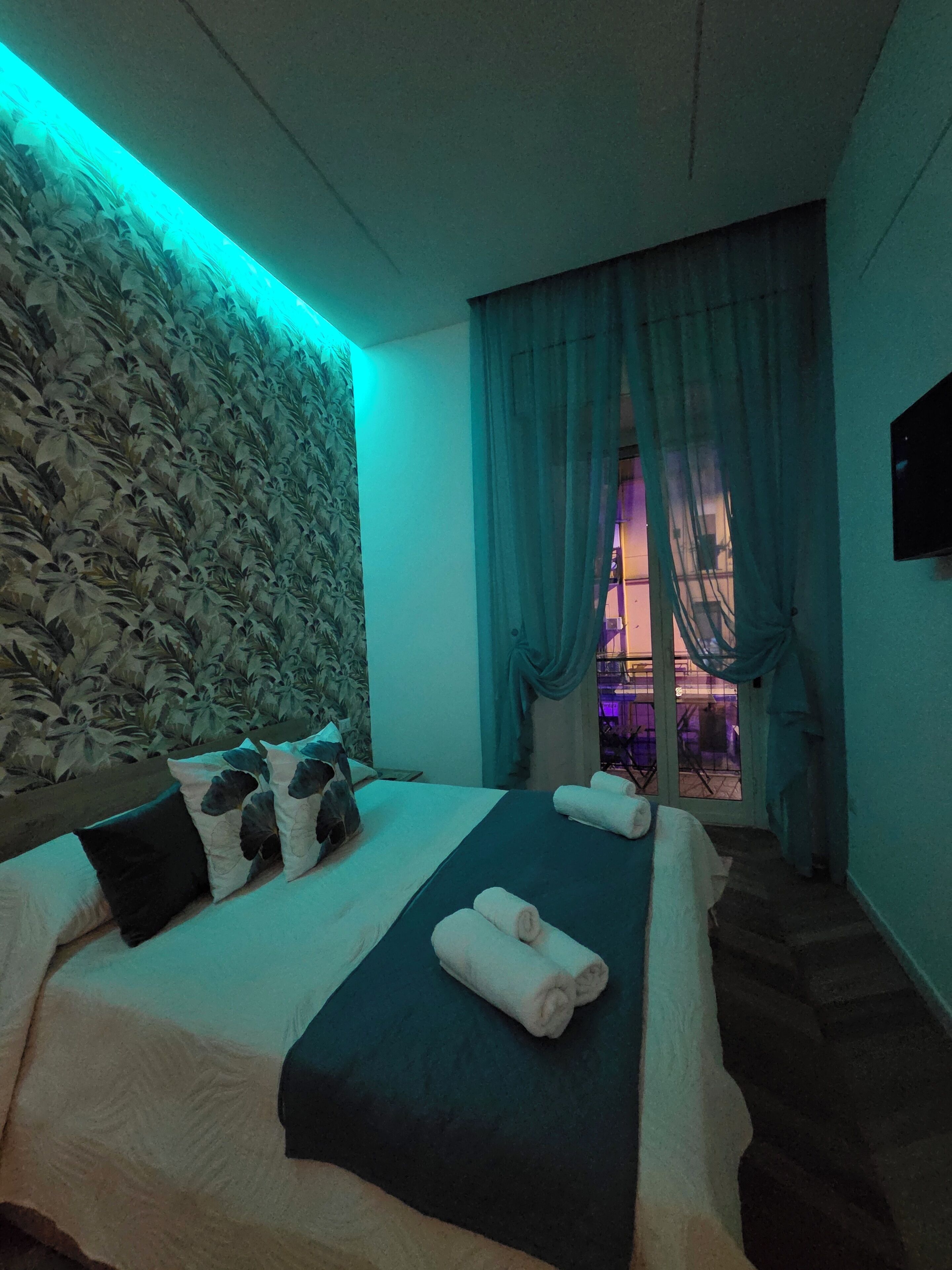 A hotel room with a bed, a wall-mounted TV, a window with curtains, and a view of the outside.