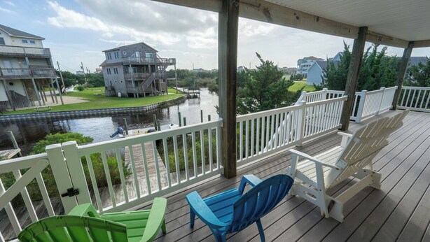 A covered deck with white railings, a green and blue Adirondack chair, and a view of a canal with houses in the background. - The Kite Club - Canal Front, All One level - 4 Bedroom House, Pool, Large Docks (Avon)