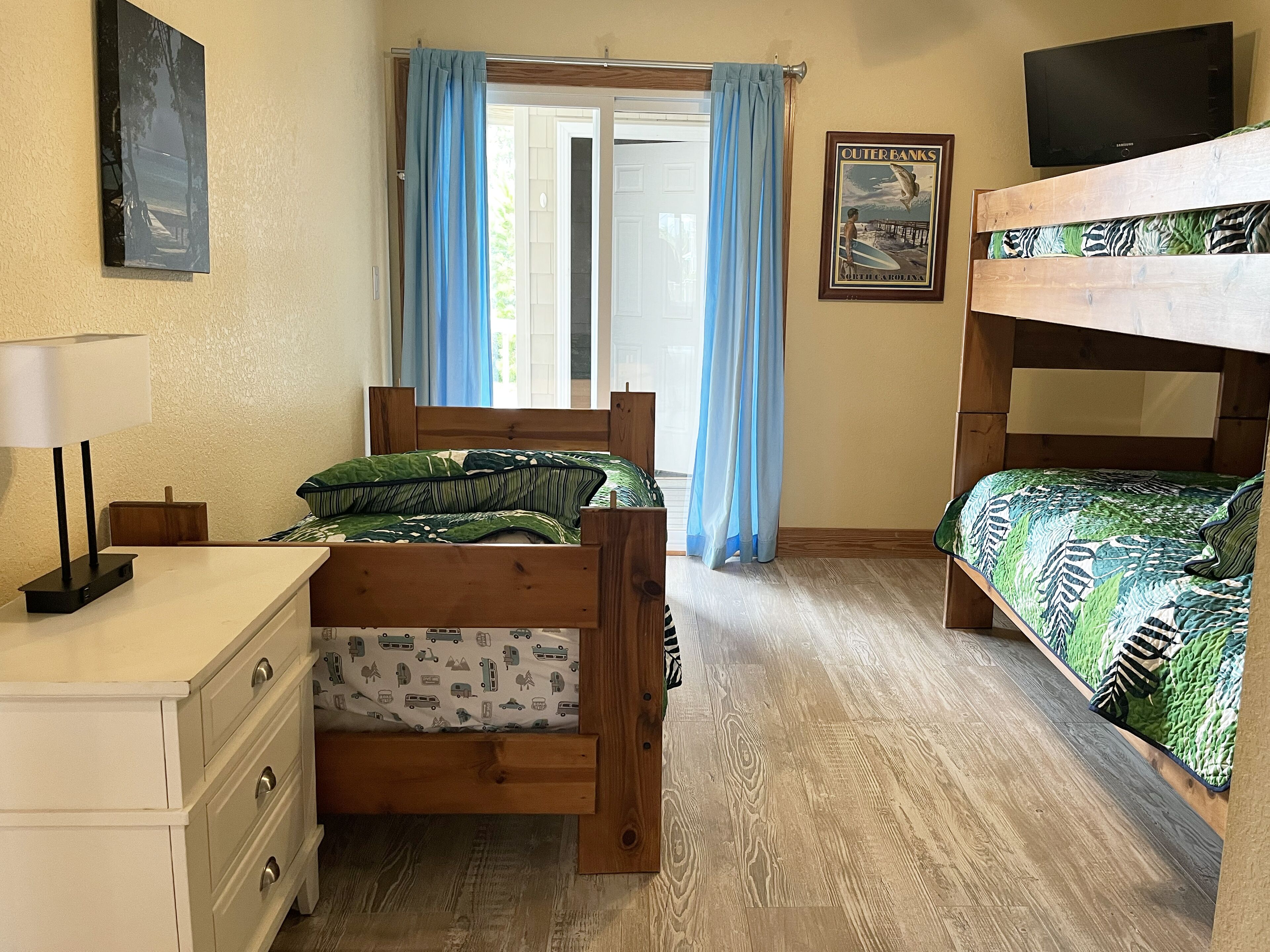 Une chambre avec un lit superposé en bois, un lit simple avec une literie à motifs, un bureau avec une lampe, un tableau encadré au mur et une fenêtre avec des rideaux.