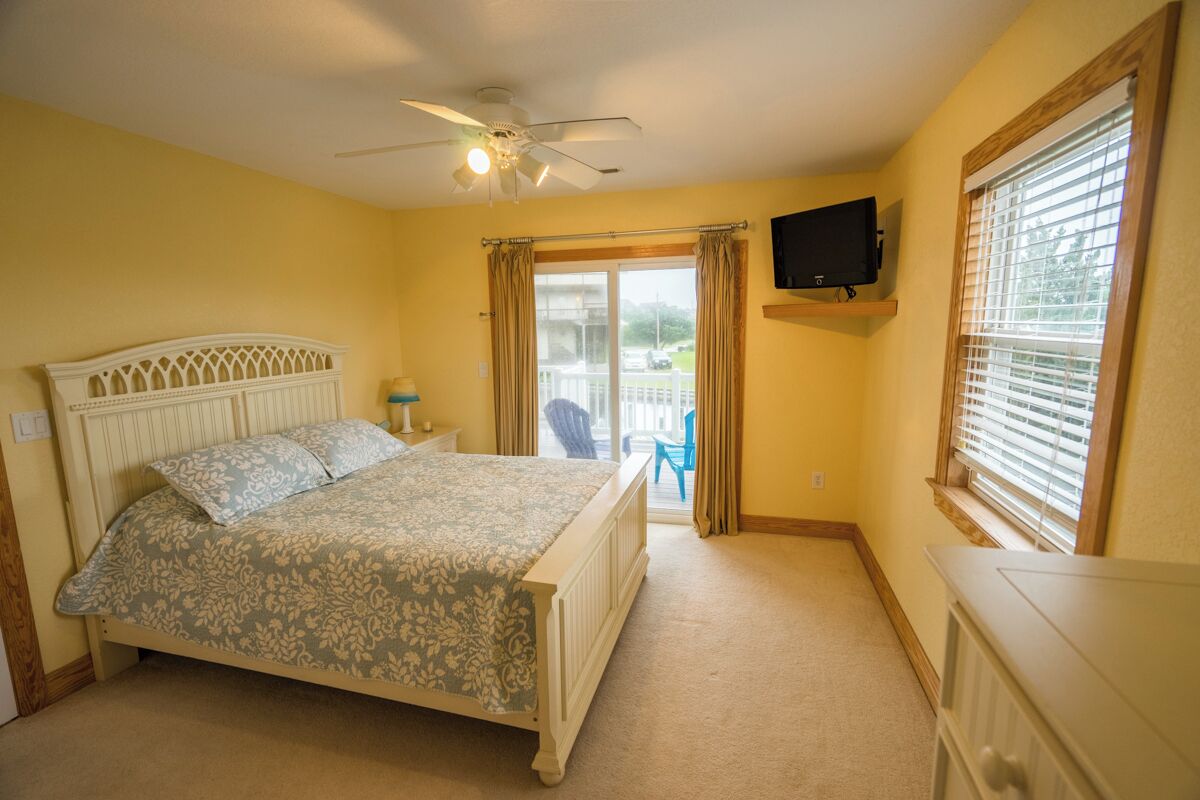 A bedroom with a bed, a ceiling fan, a television on the wall, and a window with blinds.