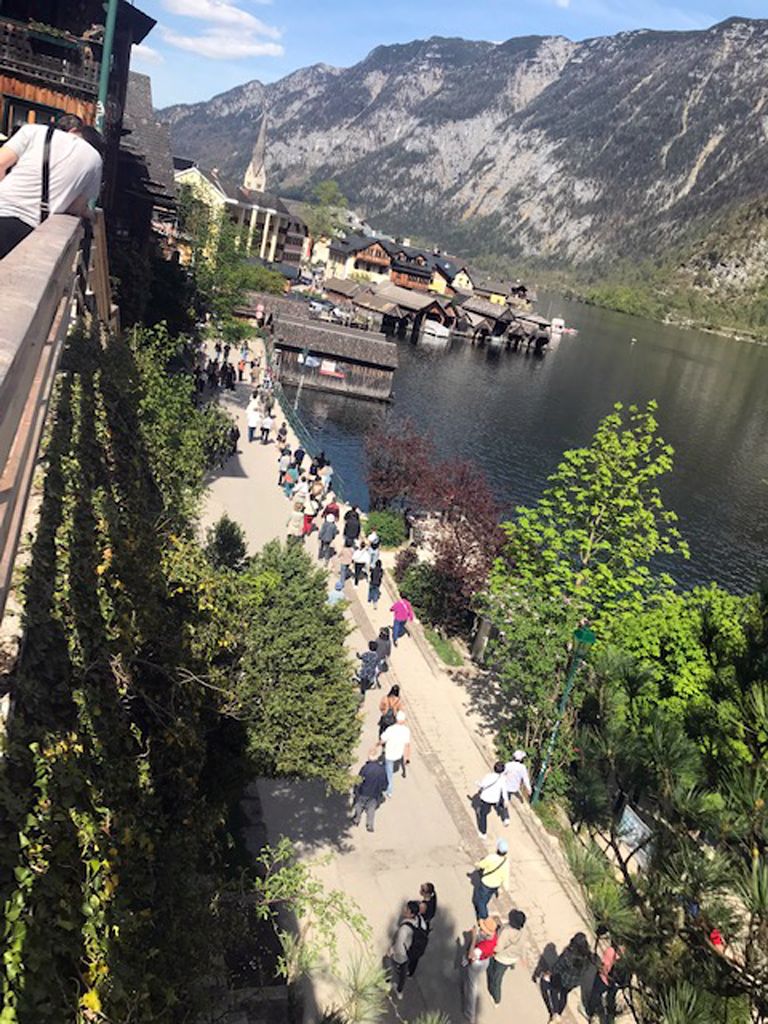 A riverside walkway with people walking, surrounded by mountains and traditional buildings.