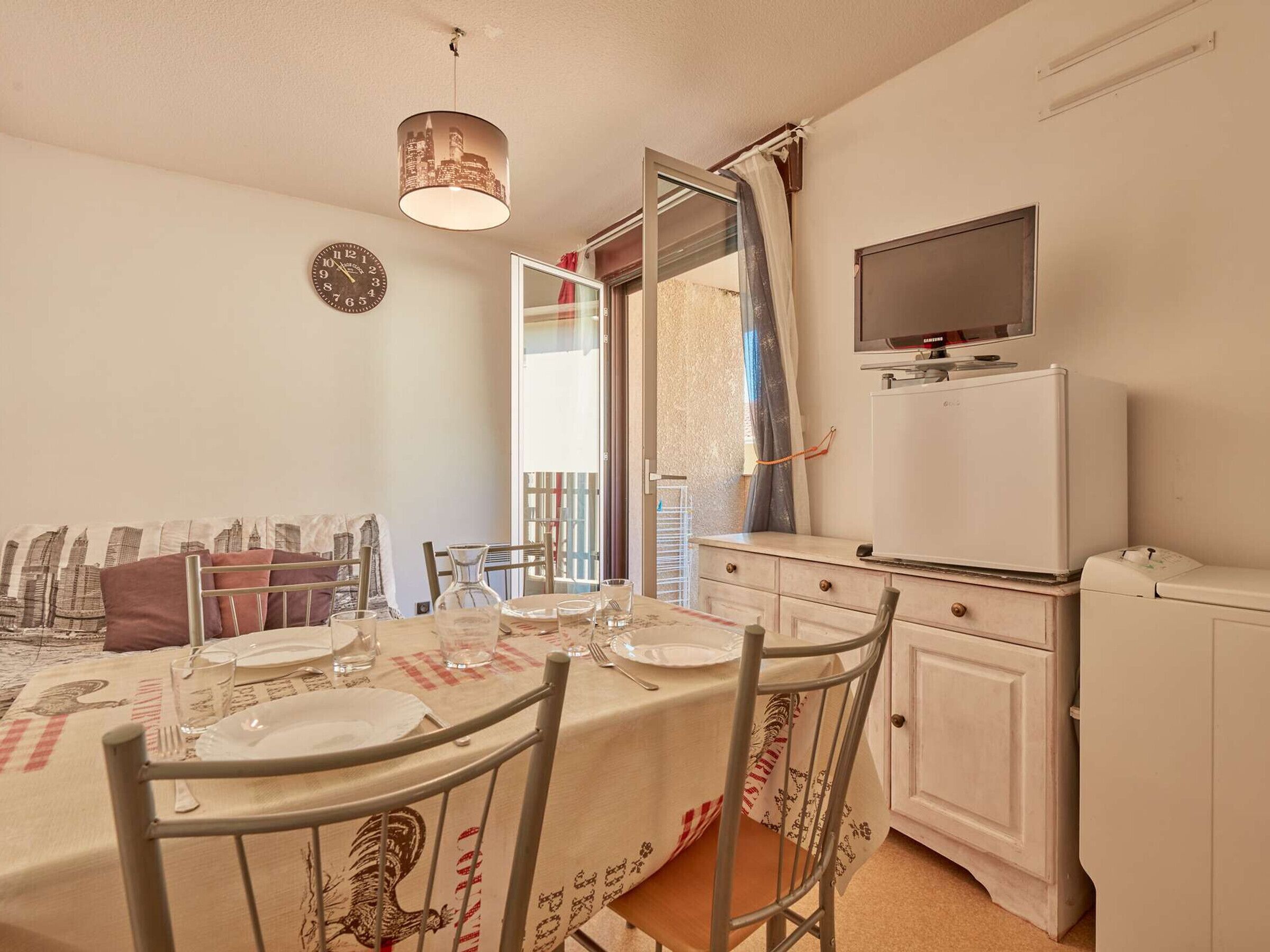 Un coin repas avec une table dressée pour quatre personnes, une horloge au mur et un téléviseur fixé au-dessus d’un meuble blanc.