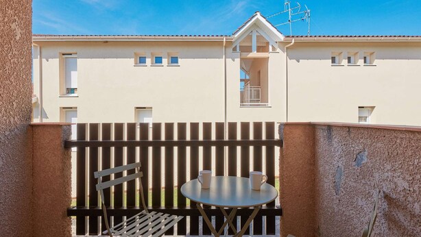 A balcony with a table and chairs, a wooden fence, and a building with windows in the background.