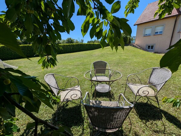 Un espace extérieur verdoyant, avec des chaises et une table en osier, entouré d’arbres et avec un bâtiment visible au loin.