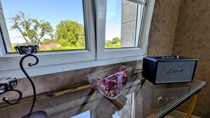 A room with a glass table, a Marshall speaker, a glass bowl with flowers, a black desk lamp, and a window with a view of greenery and buildings.