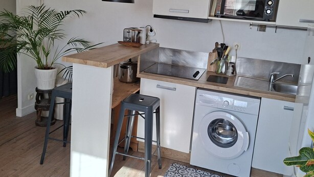 A compact kitchen with a microwave, sink, and washing machine, wooden bar counter, and two stools.