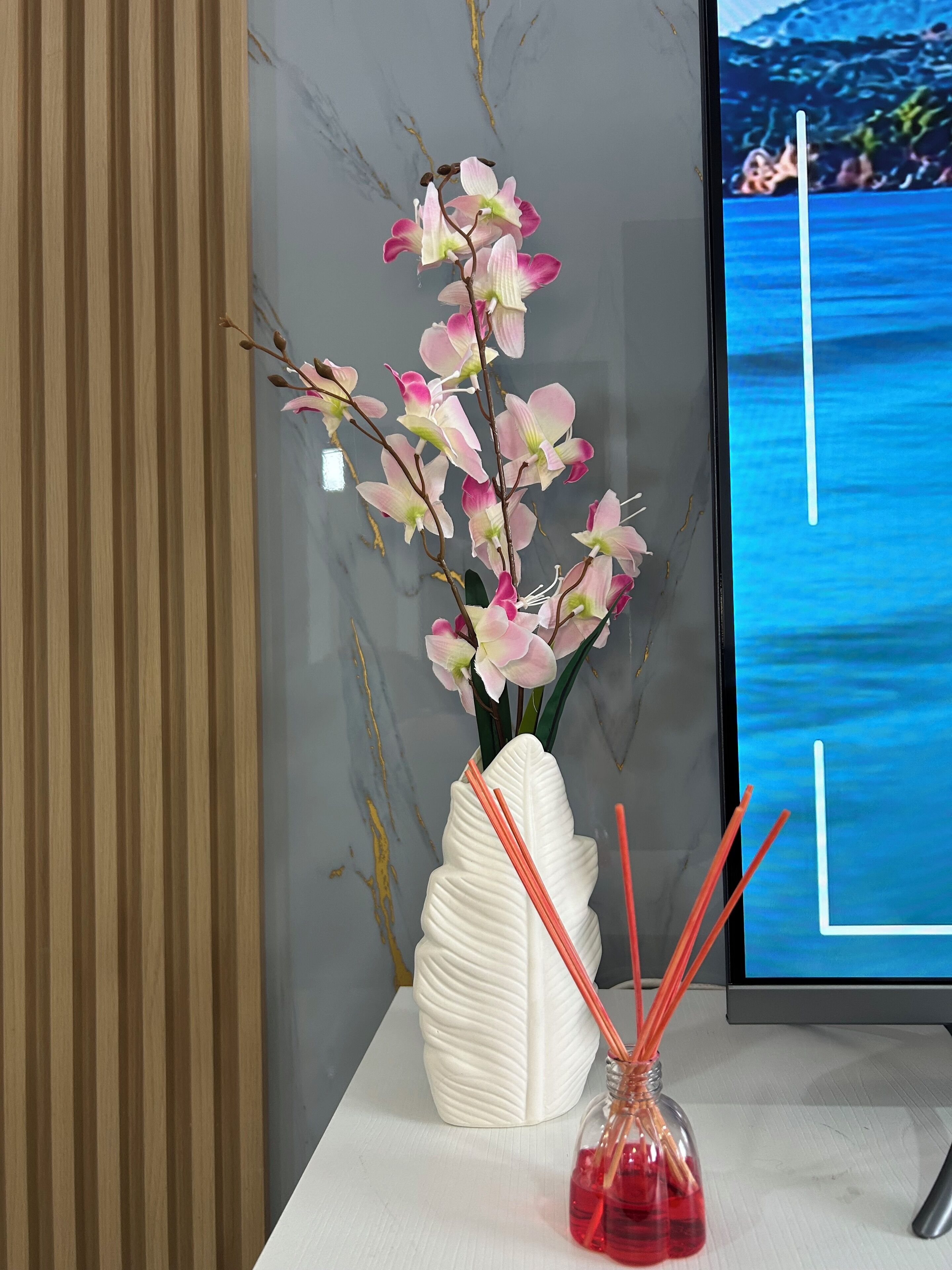Un jarrón blanco con orquídeas rosadas y cañas rojas sobre una mesa blanca, frente a una pared gris.