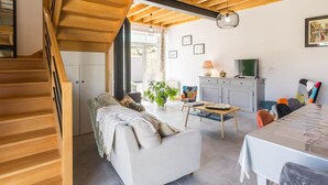 A modern living room with a staircase, wooden ceiling beams, a sofa, a dining table, and a TV. - Bright house with terrace and private garden near the beach (Bretteville)