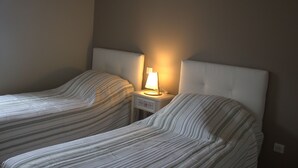 Zwei Einzelbetten mit gestreifter Bettwäsche, ein Nachttisch mit Lampe und eine schlichte Wand.