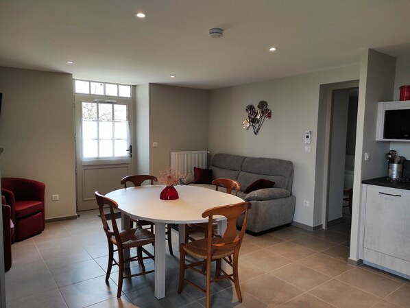 Ein Essbereich mit einem runden Tisch, Holzstühlen, einer roten Vase, einem grauen Sofa und einer Küchenzeile mit Schränken und einer Mikrowelle.