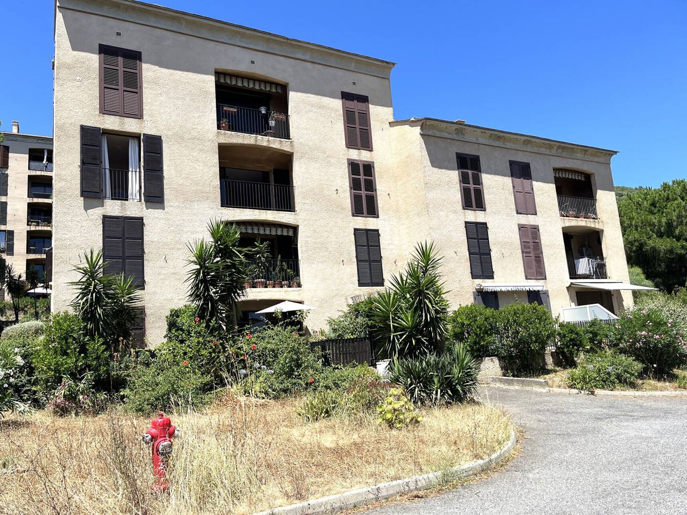 Um edifício de vários andares com varandas, rodeado por vegetação e um hidrante vermelho.