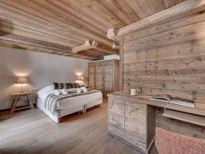 A bedroom with a wooden ceiling, a bed with white bedding, a nightstand with a lamp, and a wooden desk with a mug.