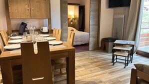 Habitación de hotel con mesa de comedor de madera dispuesta para cuatro personas, una cama, una pequeña mesa con papeles, un televisor montado en la pared y una ventana con cortinas.