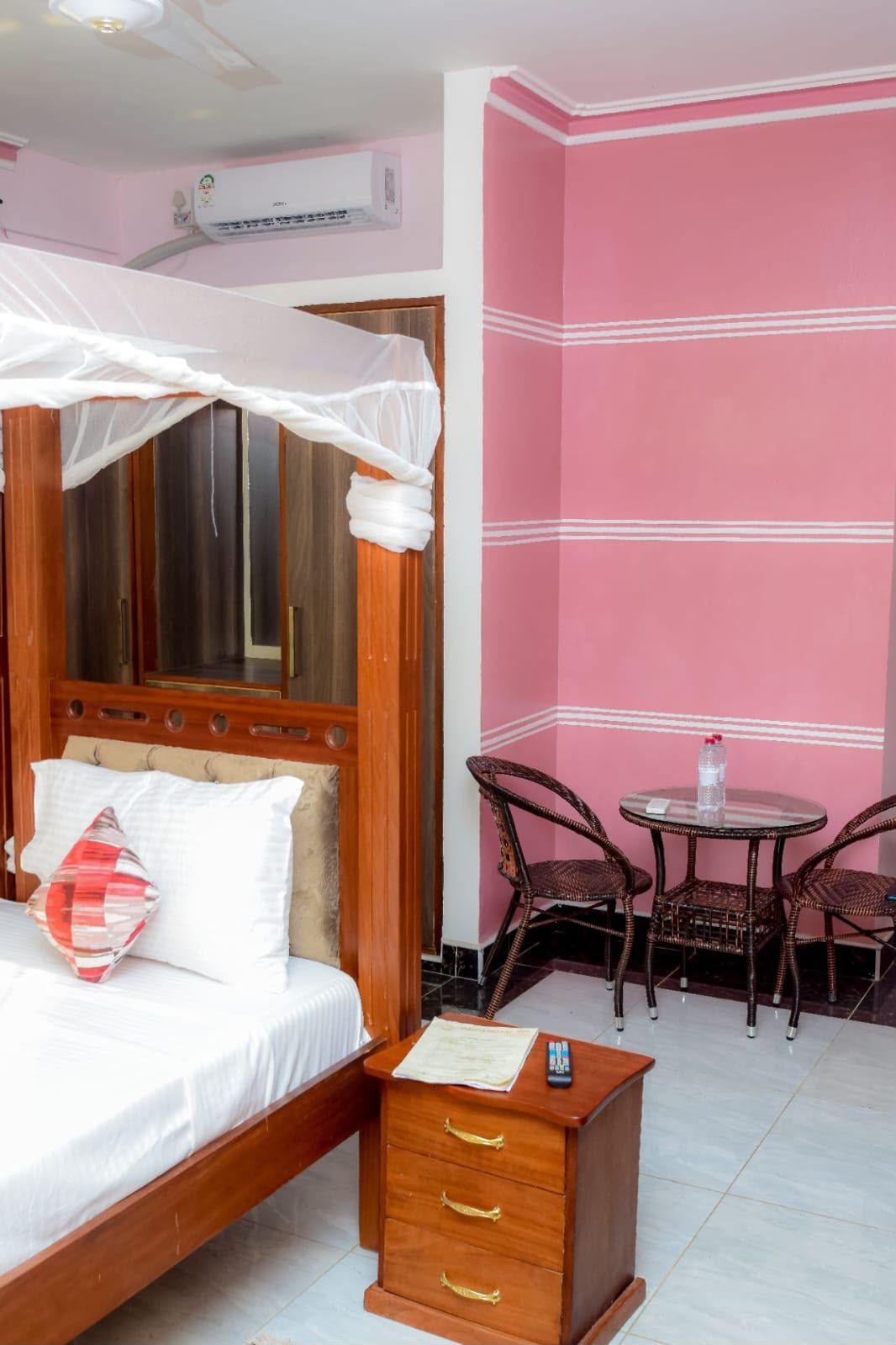 A bedroom with a wooden bed, a bedside table, a chair, a small table with a bottle, and a pink wall.