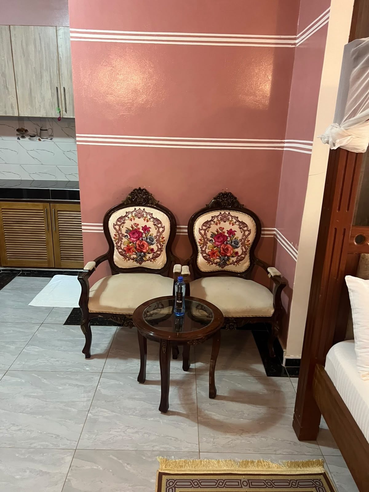 Een kamer met twee sierlijke stoelen met bloemenmotieven, een klein rond tafeltje met een fles, en een bed met een witte spreidek.