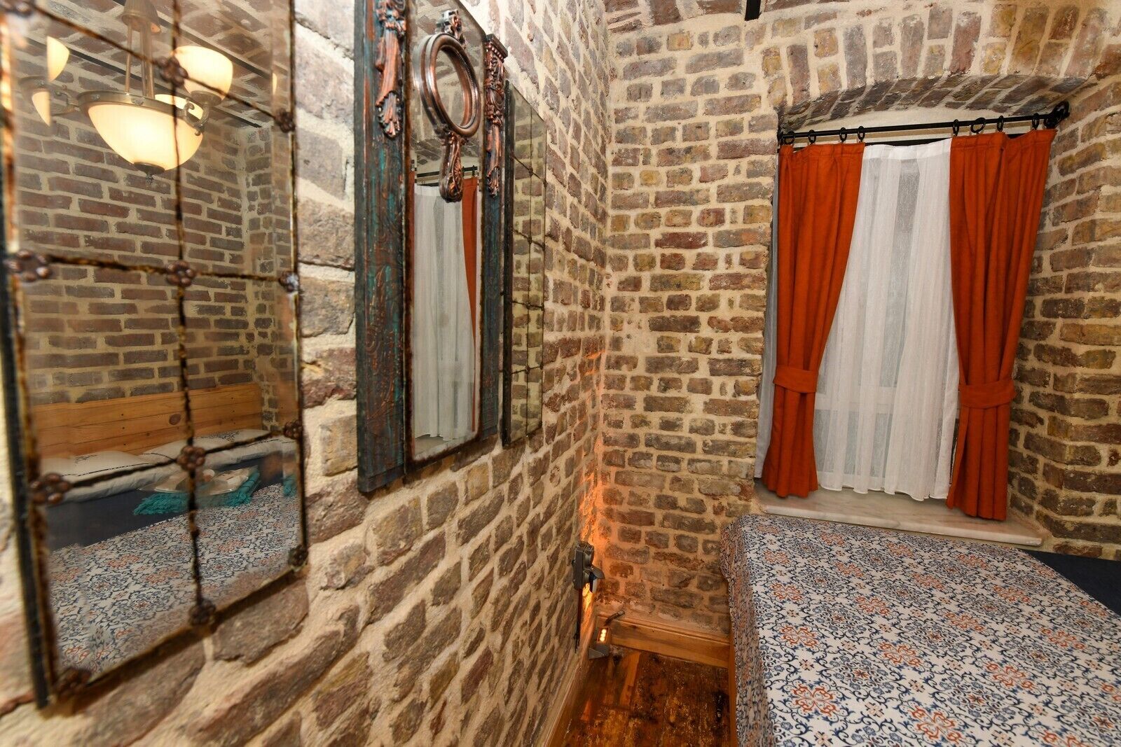 A rustic bathroom with exposed brick walls, a wooden bench, a mirror with a decorative frame, and a window with orange curtains.
