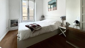 Un dormitorio minimalista con cama, mesita de noche, una silla y ventana con vistas a edificios.