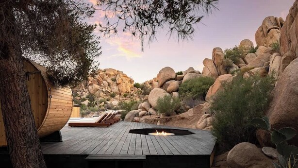 A wooden deck with a fire pit, surrounded by rocks and cacti, leading to a wooden cabin.