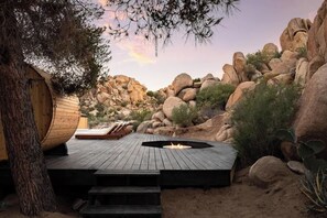 Une terrasse en bois avec un foyer, entourée de rochers et de cactus, qui mène à un chalet en bois.