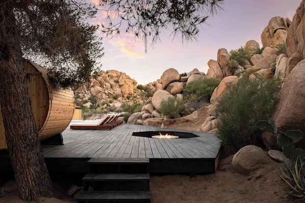 A wooden deck with a fire pit, surrounded by rocks and cacti, leading to a wooden cabin.