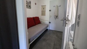 Una habitación pequeña con cama individual, colchón gris y almohadas rojas. Se ve una puerta blanca con una toalla colgada del pomo.