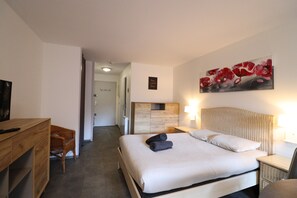 A hotel room with a bed, bedside table, a desk, a chair, a painting on the wall, and a door leading to another room.