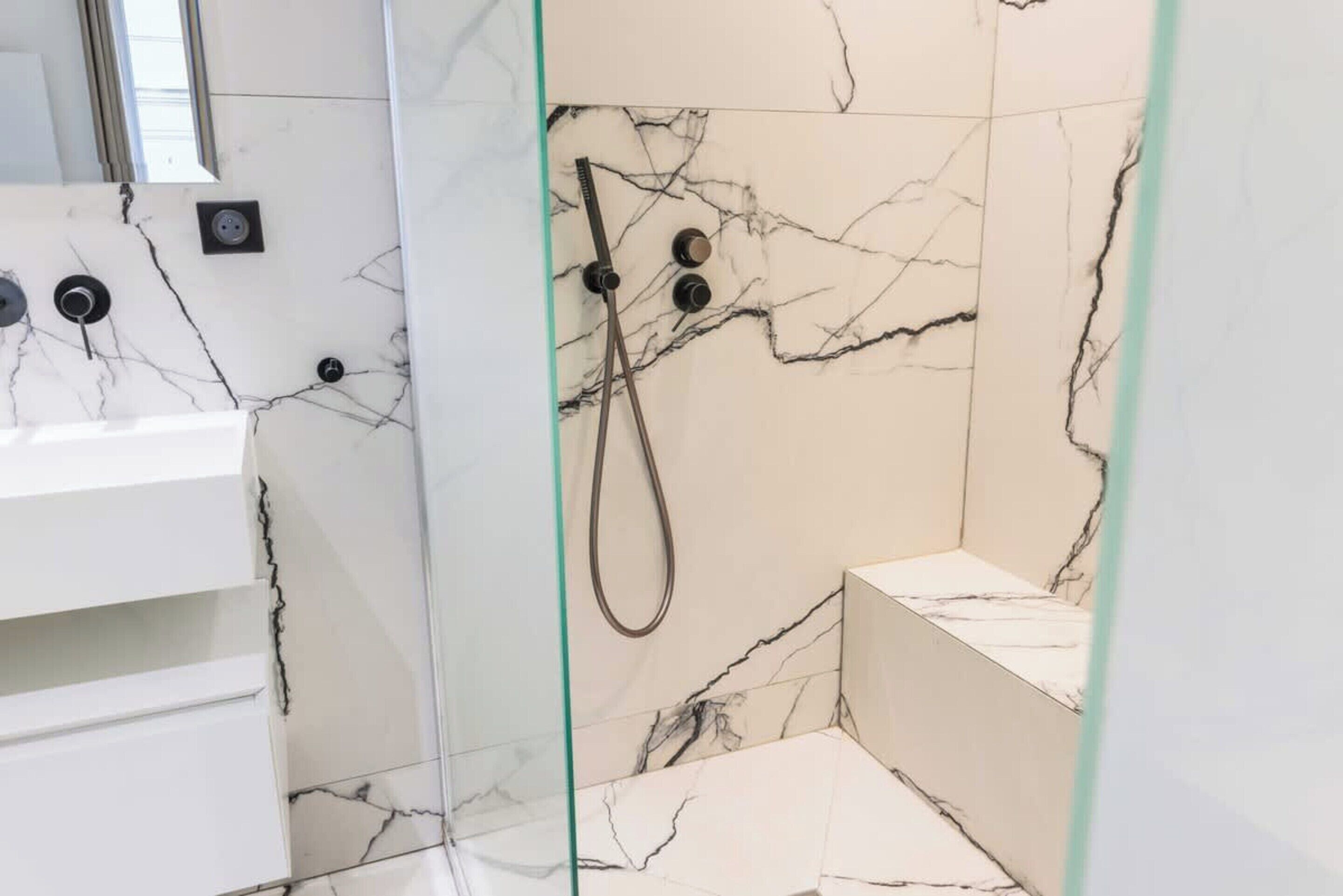 Une salle de bain avec un pare-baignoire en marbre, une douche en verre et un lavabo blanc.