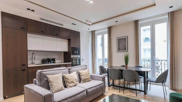 Un salon moderne avec un canapé gris, une table basse en verre, un coin repas avec une table et des chaises, et une cuisine dotée de meubles et d’appareils électroménagers aux couleurs sombres.
