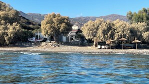 A coastal scene with a beach, a boat, and a building with a traditional roof. - NIKKON LUXURY LIVING is a luxury private villa, in top of the sea.Enjoy LUXURY. (Viannos)
